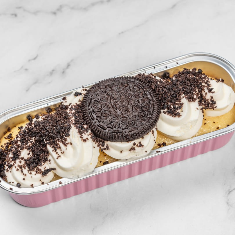 Oreo cheesecake in a pink foil pan, topped with whipped cream, cookie crumbles, and a whole Oreo.