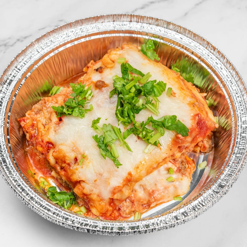 Dominican lasagna, pastelón, in a foil container, topped with melted cheese and fresh cilantro.